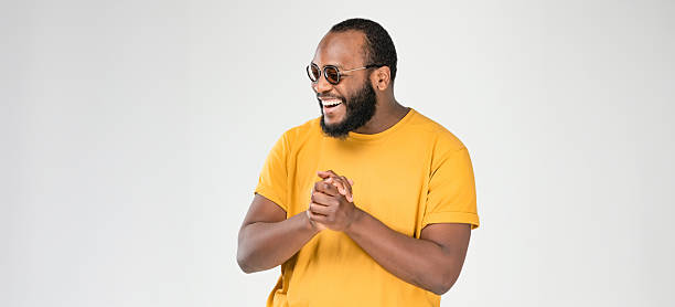 Studio portrait of a smiling man wearing a yellow shirt and glasses, clasping his hands together while looking to the side. The image represents anticipation, excitement, positive emotion and confident body language.