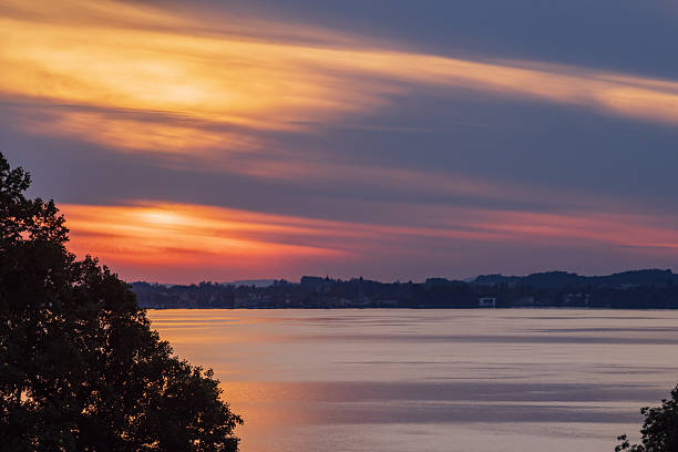 Minimalist landscape photograph of Lake Constance captured at sunset. The image shows calm water reflecting warm orange and blue tones of the evening sky, with a distant shoreline creating a balanced composition. Clean lines, soft light and natural colors emphasize tranquility and space. Suitable for travel, nature, background, relaxation and environmental concepts.