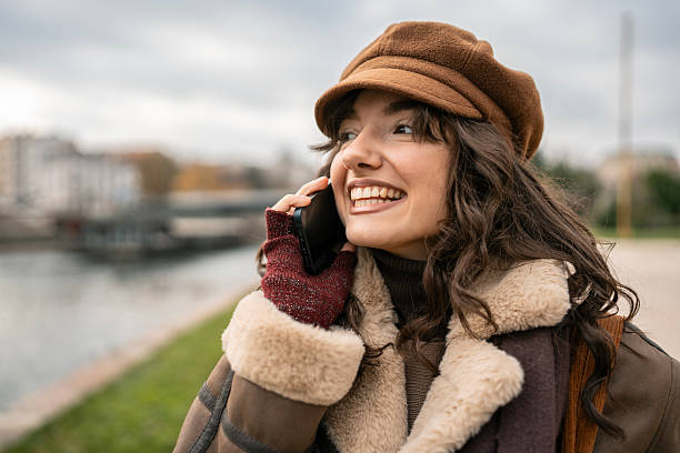 istock Joyful Woman in Brown Hat and Shearling Coat Talking on Phone by Riverfront Park 2252777675