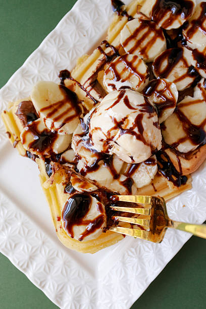 Close up top view of waffle with banana slices, vanilla ice cream and chocolate sauce. Fork lifting a bite of dessert on a white plate in bright natural light.