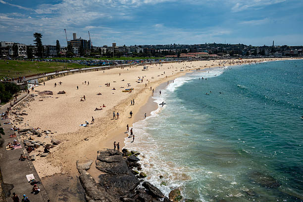 Sydney, NSW, Australia - Nov. 7, 2025: A sweeping 28mm wide-angle view from the north side of Bondi beach looking south. The historic Bondi pavilion is visible in the distance beyond the sandy shore, with surfers in the ocean and people scattered across the beach and coastal rocks
