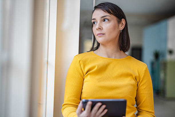 istock Woman holds tablet while standing near window in office during work hours, focusing on task and surroundings with colleagues nearby 2252801653