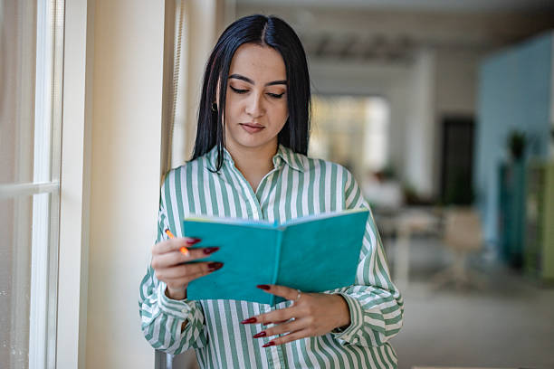 Woman in a striped shirt is standing in an office, reading a blue book. Colleagues are working in the background. The setting is modern and open during work hours.