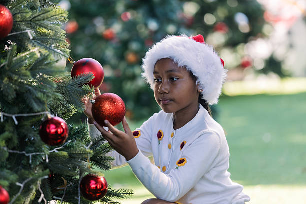 A joyful child wearing a Santa hat decorates a Christmas tree outdoors, surrounded by festive lights and ornaments. The scene captures holiday spirit, childhood happiness, family traditions, and seasonal celebration.