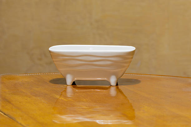 istock A white ceramic sponge holder shaped like a miniature bathtub with wavy relief patterns and four short legs, standing on a polished wooden surface against a wood-grain backdrop. 2252819395