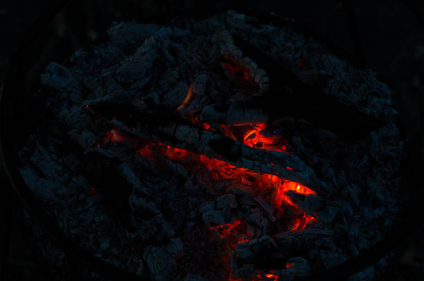 Dark scene of red-hot embers and charred coals in a fire pit. Warm, cozy glow rises from the ashes, evoking rustic evenings, camping, and intimate, ambient light.