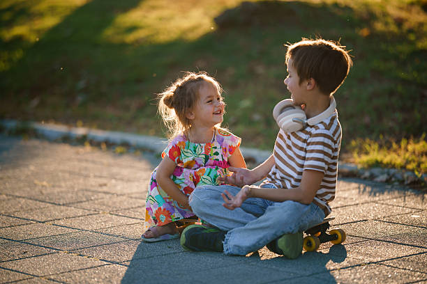 Two young children, a girl and a boy, sitting on large stone steps, engaged in conversation outdoors on a bright day.