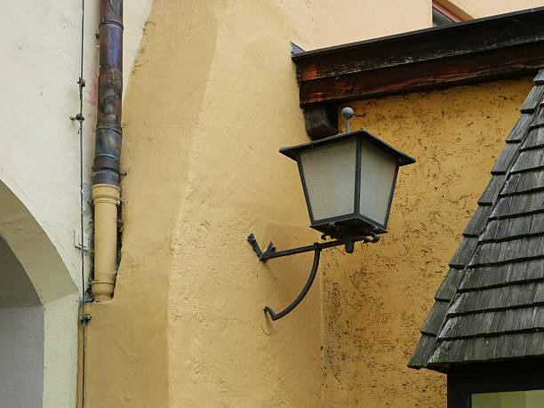 Classic wall lantern with wrought iron bracket on yellow facade with roof shingles and gutter, rustic architecture. Copyspace.