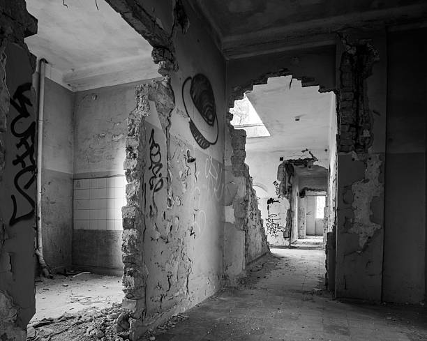 A deserted interior corridor inside an abandoned building shows crumbling walls, exposed masonry, and graffiti-covered surfaces. The black-and-white composition emphasizes decay, neglect, and the layered passage of time in forgotten urban spaces.