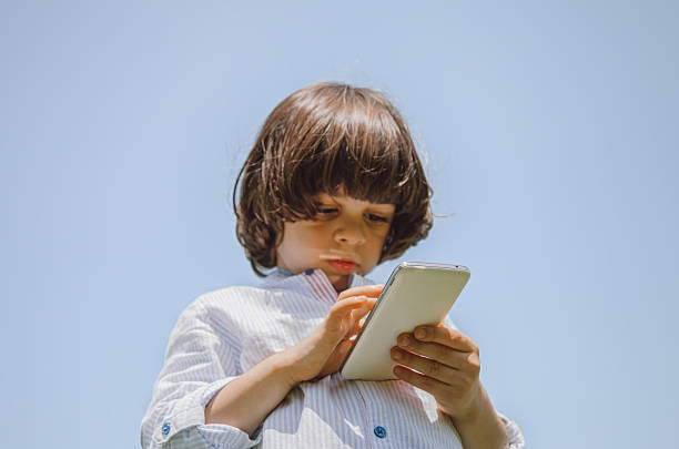 Young Arab child enjoying a sunny day outdoors while using a smartphone in a public park in Dubai, United Arab Emirates. The little boy is focused on the mobile screen, appearing to talk or interact on the phone, representing modern childhood, technology use, digital communication, and everyday family lifestyle in the Middle East. The scene reflects leisure time, warm weather, and contemporary urban life surrounded by greenery and open space.