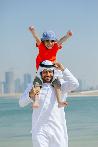 istock Smiling Arab Father Carrying Young Son on His Shoulders While Playing at the Beach with Dubai Skyline in the Background 2252835818