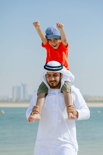 istock Smiling Arab Father Carrying Young Son on His Shoulders While Playing at the Beach with Dubai Skyline in the Background 2252835852