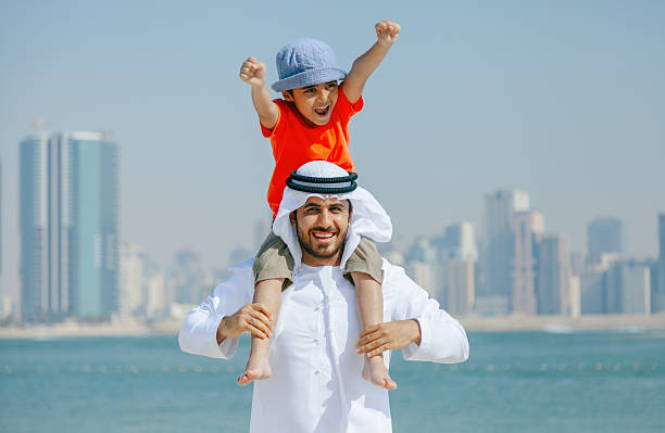 Portrait of a joyful Arab father wearing traditional white kandura carries his young son on his shoulders while playing and celebrating at a sunny beach in Dubai. The happy child, dressed in a bright orange t-shirt and blue hat, raises his arms with excitement as turquoise sea water and the modern Dubai skyline form the background. This lifestyle scene highlights fatherhood, family bonding, happiness, outdoor recreation, and Middle Eastern culture in a coastal urban setting UAE