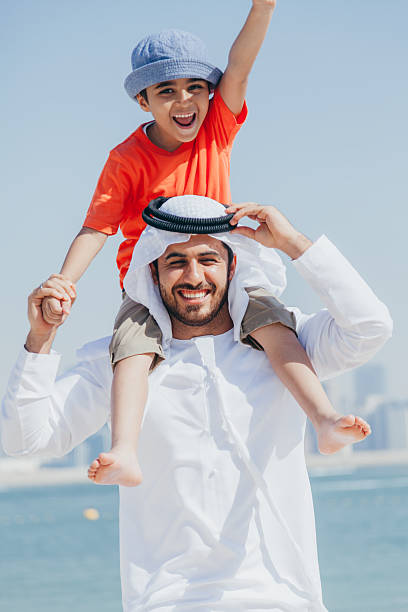 istock Smiling Arab Father Carrying Young Son on His Shoulders While Playing at the Beach with Dubai Skyline in the Background 2252835863