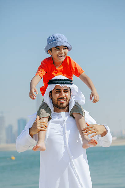 istock Smiling Arab Father Carrying Young Son on His Shoulders While Playing at the Beach with Dubai Skyline in the Background 2252835894