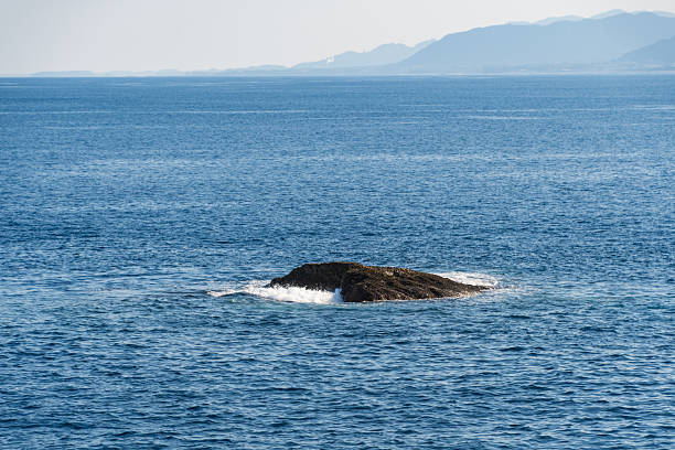 A small rock formation emerges from the vast blue ocean with white waves splashing under bright sunlight.