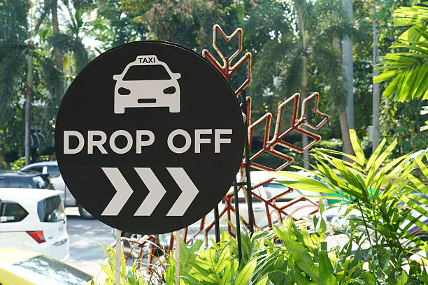 istock Round black sign with white text and graphics indicates TAXI DROP OFF with directional arrows. Surrounded by lush green tropical foliage and palm trees with parked cars in the background. 2252849845