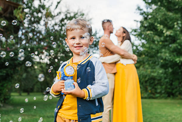 Little boy holding bubble gun with many soap bubbles floating, while mother and father hug in the background. Concept of carefree childhood, family unity and joyful moments.