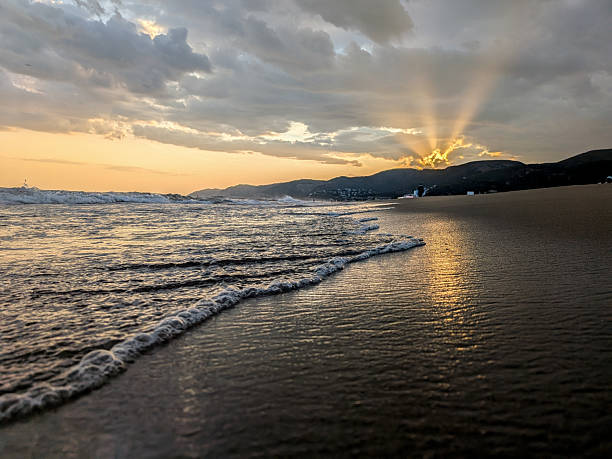 The sun setting over the distant mountains, casting dramatic sunbeams across the ocean waves as they gently roll onto the sandy beach