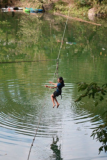 A young girl participating in a Ziplining adventure activity near a forest lake, smiling while climbing with safety harness support. The scene highlights childhood adventure, outdoor recreation, confidence building, and eco-tourism in a natural setting at Sattal, Uttarakhand, India.
