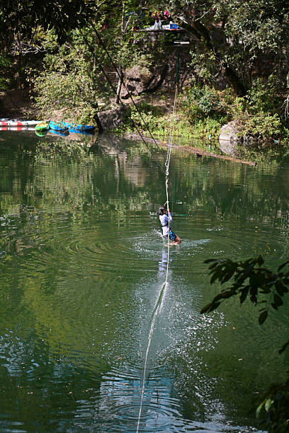 A person is balancing on a rope stretched over a canyon with water below. The scene captures the moment under clear daylight with rocks and trees in the background.