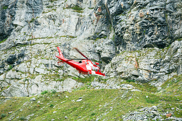 Red rescue helicopter flying low above an alpine slope with steep rocky cliffs in the background. Concept of emergency response, mountain rescue, air ambulance and helicopter operations in remote mountainous terrain.
