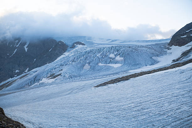 Wide view of a large alpine glacier with visible crevasses, flowing ice structure and melting zones, surrounded by rocky mountain slopes and low clouds. High altitude glacier landscape illustrating climate change, glacier retreat and global warming in the Alps