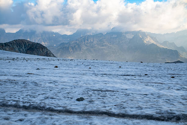 Wide view of a melting alpine glacier covered with ice, snow and scattered stones, with dramatic mountain range and clouds in the background. Concept of climate change, global warming, fragile alpine environment and high altitude landscape