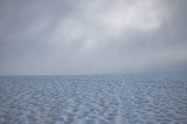 Group of alpinists moving in rope team across a wide snowy glacier and fading into dense fog. Minimalist high-altitude alpine mountaineering scene symbolizing uncertainty, risk, endurance, teamwork and extreme mountain conditions