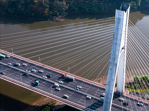 This aerial photograph captures a bustling bridge with numerous vehicles traversing it, set against a backdrop of a wide river and lush greenery. The image showcases urban infrastructure and transportation dynamics, offering insights into city planning and traffic flow. Its visual style is realistic and detailed, making it suitable for use in articles about infrastructure development, urban studies, or transportation systems.