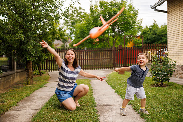 Mother and toddler son having fun playing in the backyard of their home, throwing toy planes