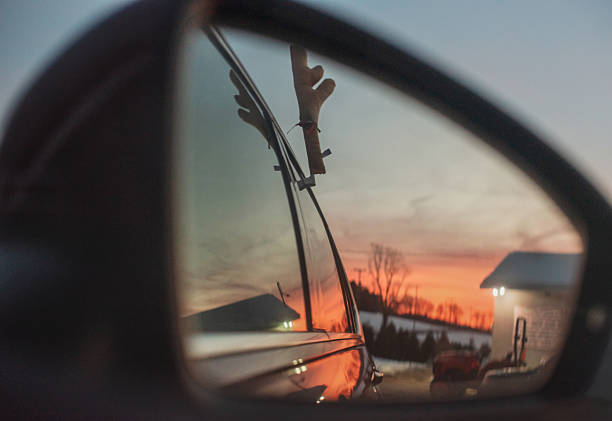 A close-up of a car's side-view mirror reflecting a vibrant winter sunset. A novelty reindeer antler decoration is visible on the car's window in the reflection. The background shows silhouetted trees and a building against an orange and blue sky. This shot uses selective focus, with the reflection sharp and the mirror's frame blurred.