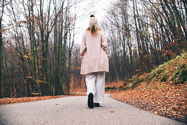 A woman in a soft pink coat and beanie walks down a winding forest road in autumn, surrounded by colorful fall foliage and a calm, contemplative mood.