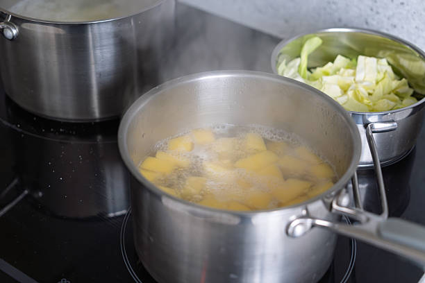 A close-up view of two stainless steel pots on a black induction stovetop in a contemporary kitchen. In the foreground, a pot filled with chunks of yellow potatoes boiling in water, with visible steam rising from the surface. Adjacent to it, another pot contains freshly chopped green cabbage leaves. The scene captures the active cooking process of preparing vegetables, emphasizing the simmering heat and bubbling water. No people are present. This image suits commercial uses such as recipe illustrations, cooking tutorials, kitchen appliance promotions, food preparation guides, and lifestyle content focused on healthy home cooking or meal prep routines.