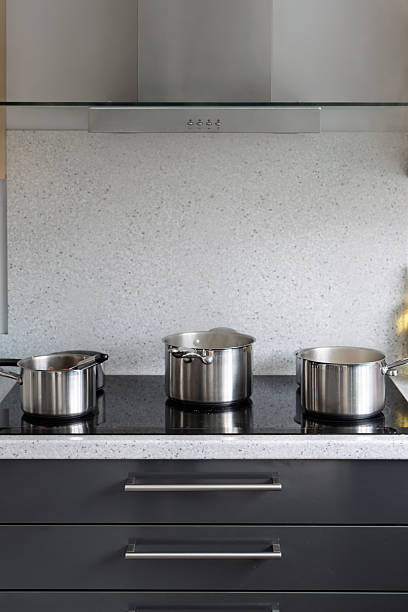 Three stainless steel pots of varying sizes sit on a sleek black induction cooktop, ready for cooking. The setup features a glossy white speckled backsplash, a glass range hood overhead, and dark wooden cabinets below. This clean, minimalist image captures a contemporary kitchen environment with metallic silver tones dominating the scene. Ideal for commercial use in articles on home appliances, cooking tutorials, interior design features, or product catalogs showcasing modern kitchenware and induction technology. The composition highlights functionality and hygiene in domestic settings, suitable for media content on healthy cooking or home renovation trends.