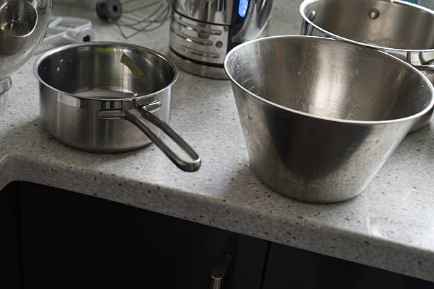 A collection of shiny stainless steel cookware including saucepans with handles, large mixing bowls, and a stand mixer sits on a light gray quartz countertop in a contemporary kitchen. Dark wooden cabinets are visible below the counter, with a white towel and metal whisk nearby. The setup appears organized and ready for meal preparation, conveying a clean and efficient cooking environment. This image suits commercial applications such as recipe illustrations in cookbooks, promotional materials for kitchen appliances and utensils, lifestyle articles on home cooking, and advertising for modern kitchenware brands targeting home chefs and food media.