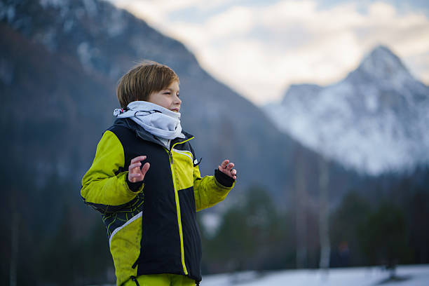 Boy wearing a winter jacket and scarf standing outdoors in a snowy mountain landscape.