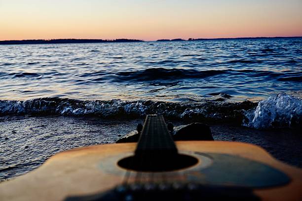 An acoustic guitar rests near waves and ripples along a shoreline at sunset; this scenic capture evokes emotional expressions such as of freedom, creativity, inspiration