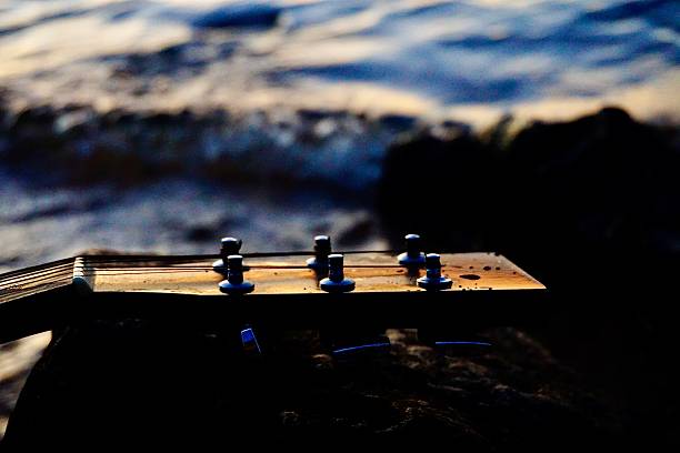 A close-up of tuning knobs of an acoustic guitar rests near waves and ripples along a shoreline at sunset; this scenic capture evokes emotional expressions such as of freedom, creativity, inspiration