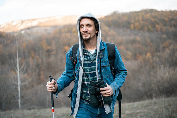 A rugged hiker in a denim jacket with a hoodie, carrying binoculars and a walking stick, trekking through a fall forest. Perfect for adventure, exploration, outdoors, and travel themes.