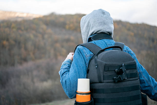 A lone hiker wearing a gray hoodie and denim jacket carries a black tactical backpack along a mountain trail, with autumn forest in the background, conveying exploration, adventure, and outdoor fitness.