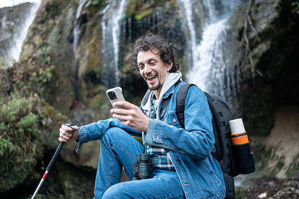 istock Smiling Hiker With Backpack Using Smartphone By Waterfall On Outdoor Adventure Trek 2252890908