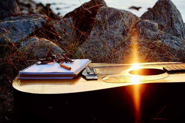 A notepad and pencil sits on top of a guitar during a sunset as sun rays shine on the guitar strings.