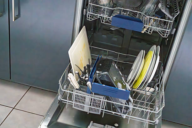A close-up view of an open dishwasher in a contemporary kitchen, with the lower rack pulled out and filled with clean white plates, bowls, and stacked kitchen utensils including blue-handled tongs and silverware. The upper rack holds additional dishes and cups. Surrounding elements include a stainless steel sink with a yellow sponge, white quartz countertops, gray matte cabinets, stainless steel pots and pans on the counter, and a tiled floor. The scene conveys a sense of organized household chores and modern domestic efficiency. Ideal for commercial use in articles on home appliances, kitchen organization, cleaning routines, interior design for residential spaces, and lifestyle content about everyday home maintenance by authors and media companies.