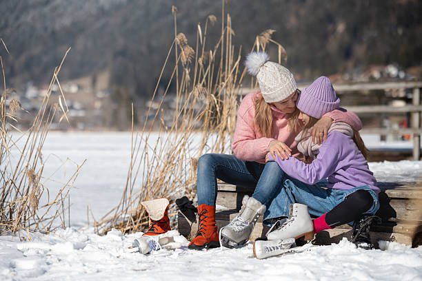 During a winter school holiday trip, a mother and daughter sit on the steps of a lakeside hut on a natural ice rink Weissensee in the Austrian Alps, getting ready to ice skate, the mother hugs her daughter with maternal love.