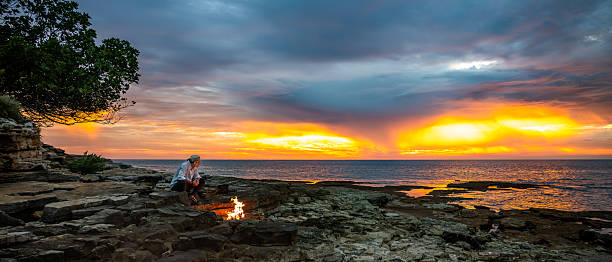 A lone person sits by a campfire on rocky shores at sunset, embracing solitude and mindfulness. The vibrant sky and calm sea create a serene, introspective atmosphere, ideal for mental wellbeing and escaping daily life.