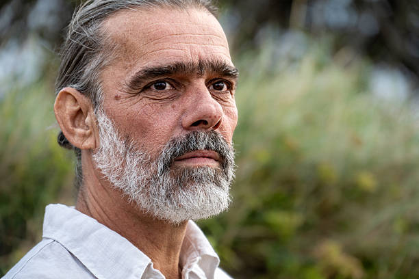 An older man with a gray beard stands in nature, exuding mindfulness and mental well-being. His focused gaze and serene expression suggest introspection, capturing a moment of getting away from it all.