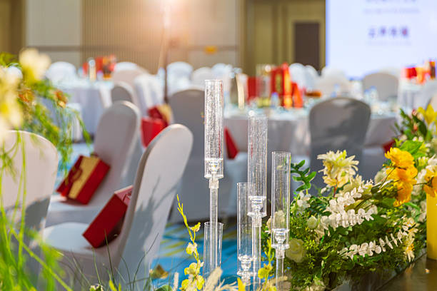 A close-up of clear tall candle holders and lush floral arrangements in yellow and white that run along the table. In the blurred background are round banquet tables with white chair covers and gift bags, warm lighting producing a soft glow. The shallow depth of field emphasizes the texture and colors of the centerpiece while conveying an elegant, festive mood.