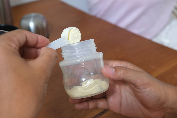 Close-up POV shot of unrecognizable hands scooping powdered infant formula from a small plastic container using a measuring scoop.Concept: baby formula, infant milk powder, powdered formula, baby nutrition, infant feeding, formula preparation, childcare, newborn care, parenting, baby food, unrecognizable hands