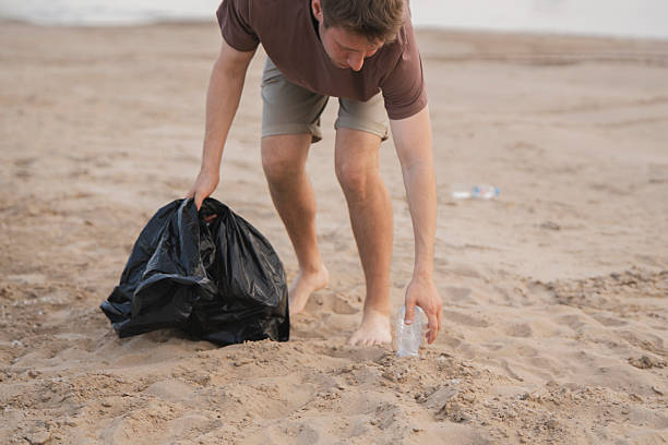 istock Man collects trash in a black bag while cleaning a sandy outdoor area 2252907680